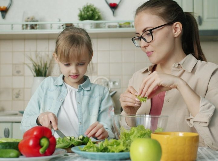 Cute loving daughter is helping mother with salad cutting cucumbers feeding young woman and laughing having fun at home. People and cookery concept.
